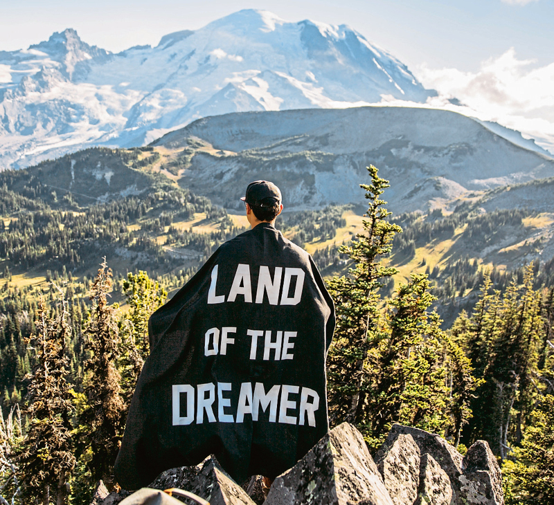 Person mit Umhang, auf dem "Land of the Dreamer" steht, schaut auf eine Berglandschaft