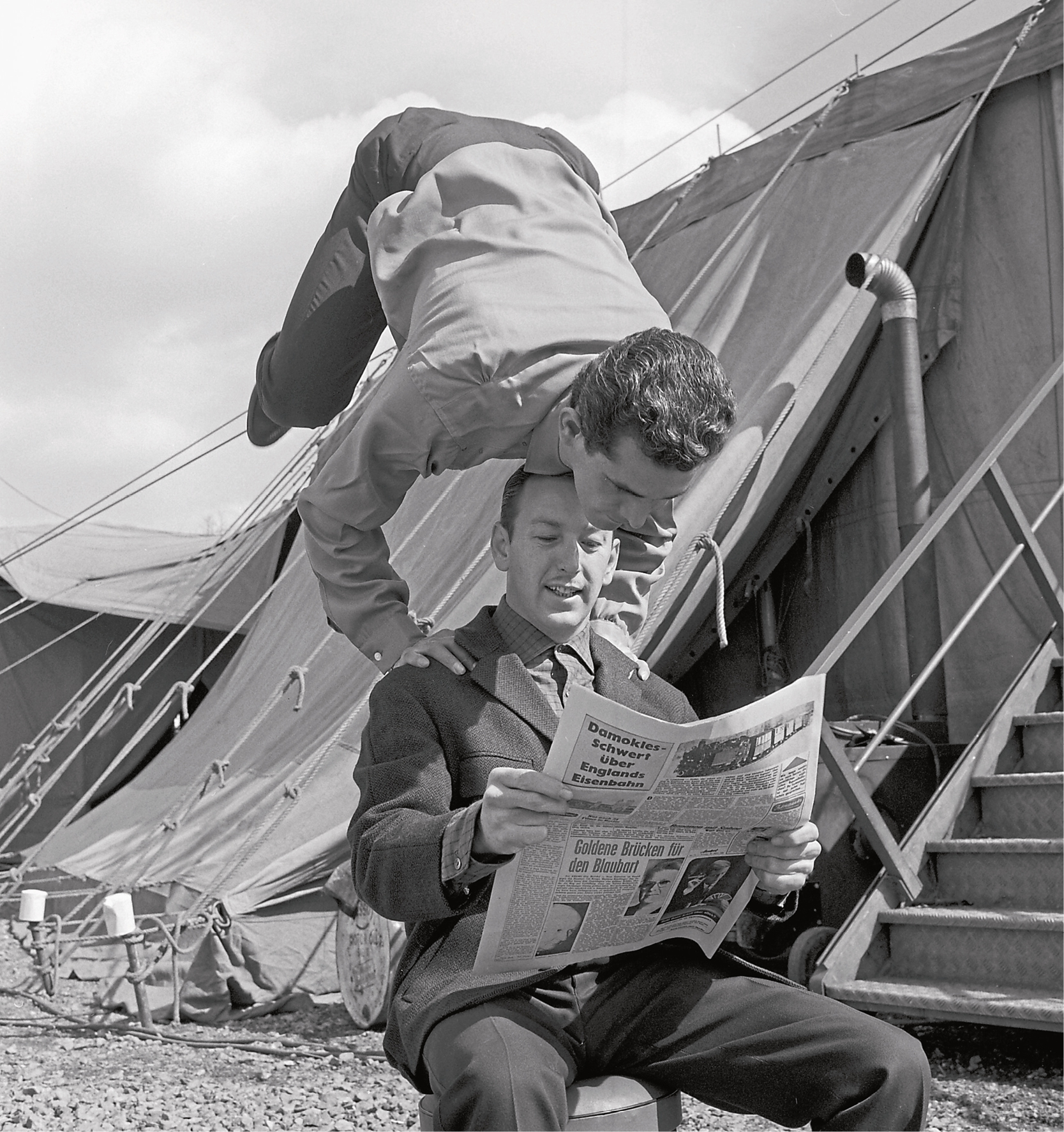 Ein Mann stützt sich auf einen anderen Mann und hebt dabei die Beine in die Luft, während beide Zeitung lesen